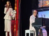A woman sings into a microphone while a man plays a black grand piano on stage, both dressed in light-colored suits.
