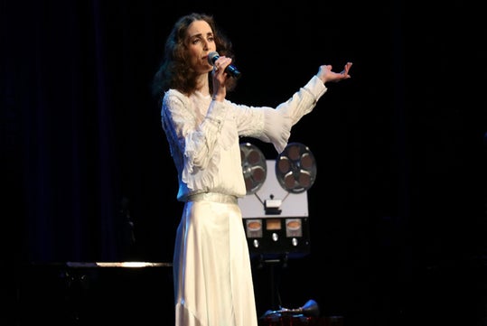 A woman in a white blouse and skirt sings into a microphone on stage, with a reel-to-reel tape machine in the background.