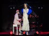 A woman in a long white dress sings into a microphone while a man in a light-colored suit plays piano onstage under bright lights.