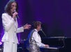 A woman in a white suit sings into a microphone while a person plays a grand piano on stage, with purple lighting in the background.