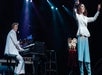 Two performers in white suits on stage; one plays the piano and sings, while the other stands with arms raised. Stage lights and musical equipment are visible in the background.