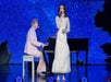 A man plays piano while a woman in a white dress sings on stage, with a blue starry night backdrop behind them.