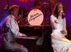 Two musicians perform on stage; a man plays the piano while a woman sings into a microphone. A drum in the background displays the name "Karen Carpenter.