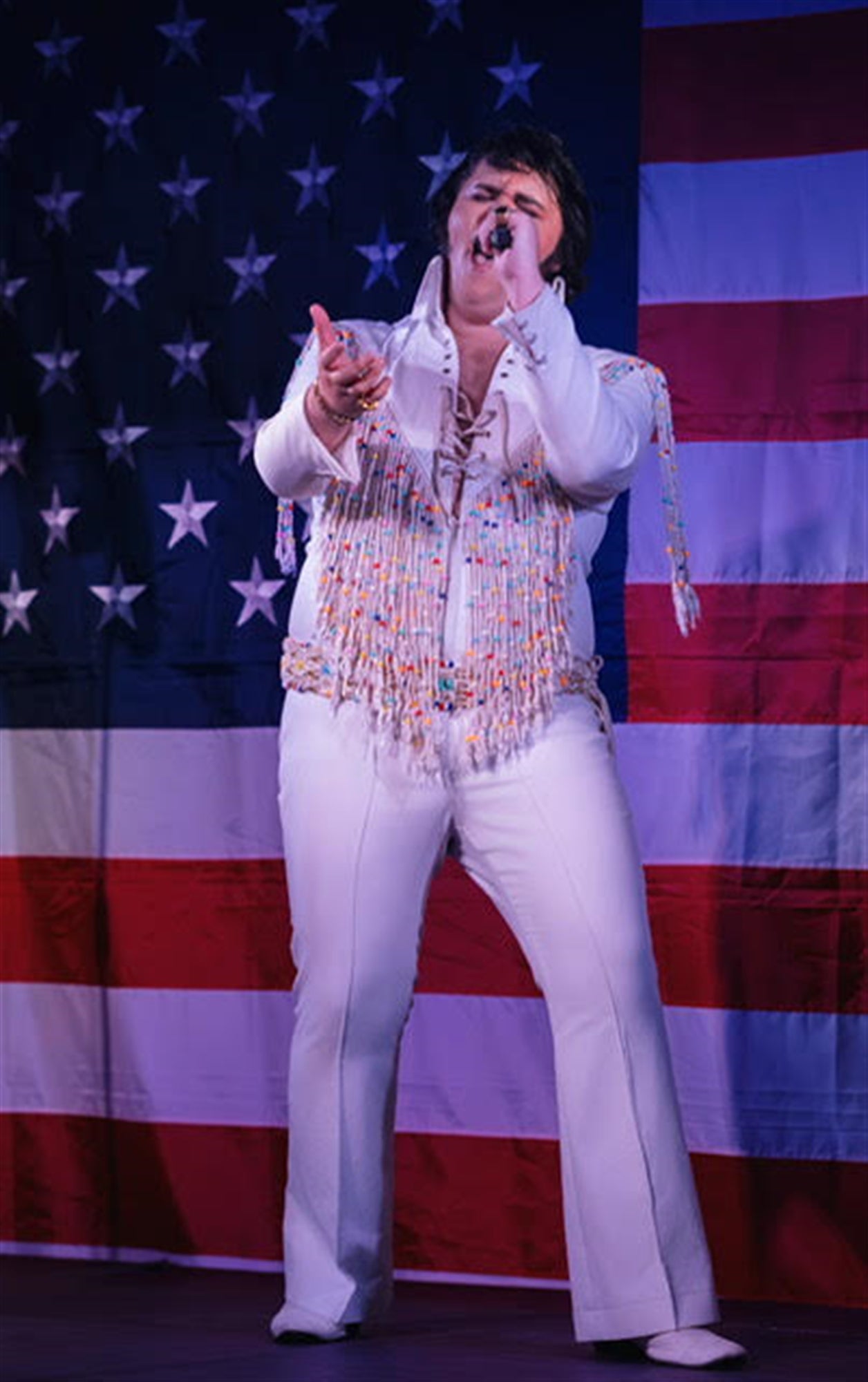 A person dressed in a white, beaded jumpsuit sings into a microphone in front of a large American flag backdrop.