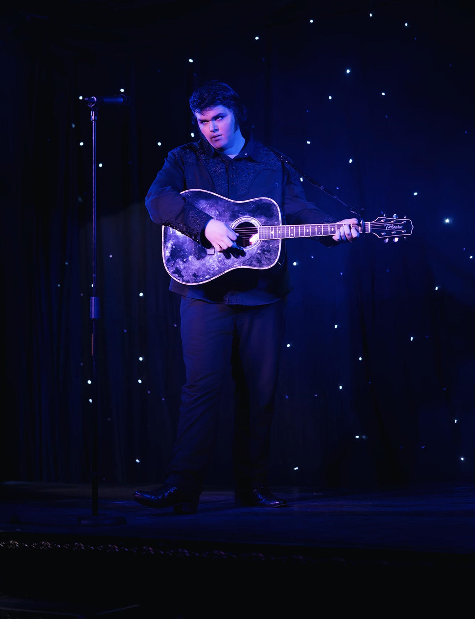 A person stands on a stage playing an acoustic guitar under blue lighting, with a microphone stand nearby and a backdrop of small, glowing lights.