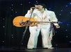 A person dressed in a white jumpsuit with fringe and holding a guitar stands on stage with a microphone, in front of a backdrop with small blue lights.