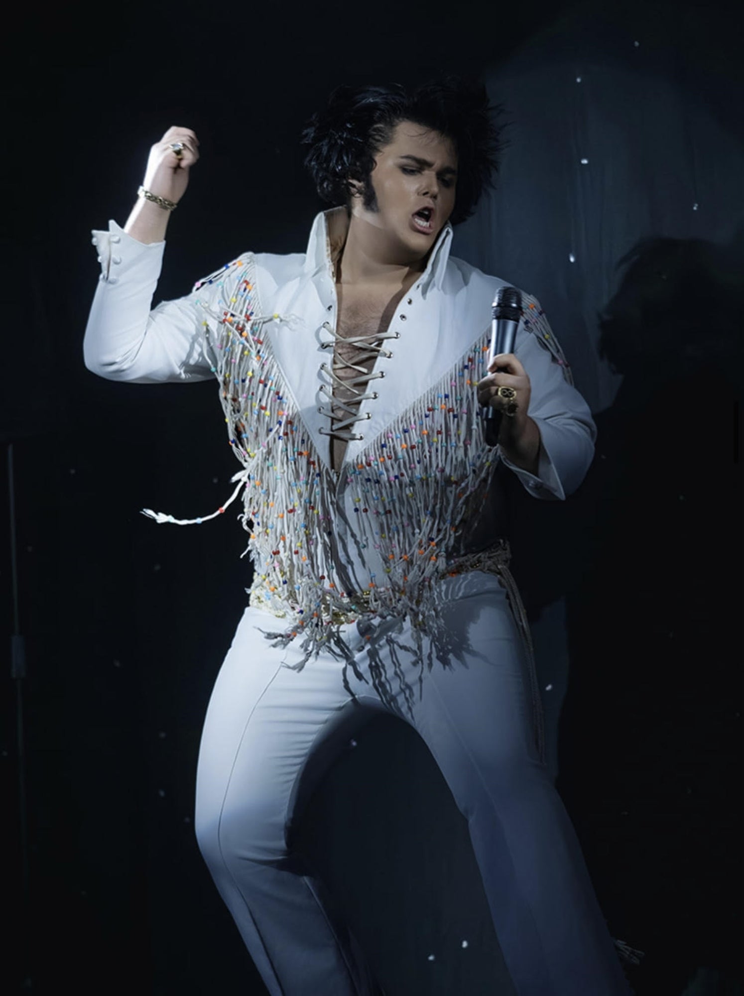 Person in a white fringe jumpsuit performs energetically on stage, holding a microphone, with dramatic lighting and a dark background.