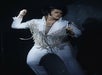 Person in a white fringe jumpsuit performs energetically on stage, holding a microphone, with dramatic lighting and a dark background.