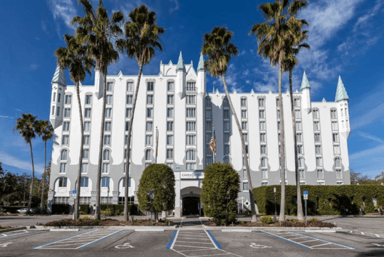 View of the bright white front of the Castle Hotel from the parking lot with tall shrubs in front of it and a blue sky overhead.