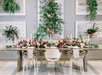 A long table with white and clear chairs at it and a floral piece with lots of pink flowers and greenery with candles mixed in.