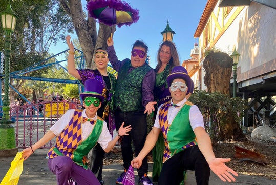 Five people in colorful Mardi Gras costumes and masks pose energetically on a sidewalk lined with trees and buildings.