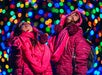 Two children in pink winter clothing look up, surrounded by colorful, blurred holiday lights.