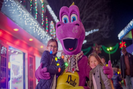 Two children pose with a purple dinosaur mascot in front of festive holiday lights at night.