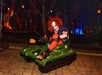 A clown with wild red hair and exaggerated makeup, sitting in a toy tank at night at Castle Park.