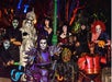 A group of people in elaborate Halloween costumes, including skeletons, a ghostly figure, and a character with a pumpkin, posing together in a colorful, illuminated outdoor setting.
