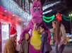 Adults and children interact with a person in a purple dinosaur costume, surrounded by holiday lights at night.