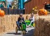 Two children ride green toy tractors through a path lined with hay bales and large pumpkin decorations at an outdoor autumn festival.