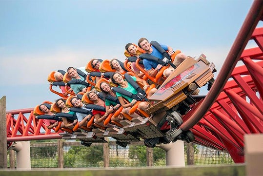 A roller coaster car full of people strapped in laughing while going around a curve on a red track on a sunny day.