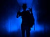 A person in a top hat with long, claw-like fingers is silhouetted against blue light in a dark hallway.