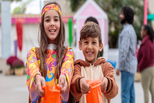 Two children in costumes hold out orange bags, smiling at an outdoor event with other people and colorful decorations in the background.