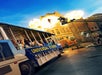 A Universal Studios tour tram passes by a film set with a stunt performer propelled by an explosion and a camera crane capturing the action.