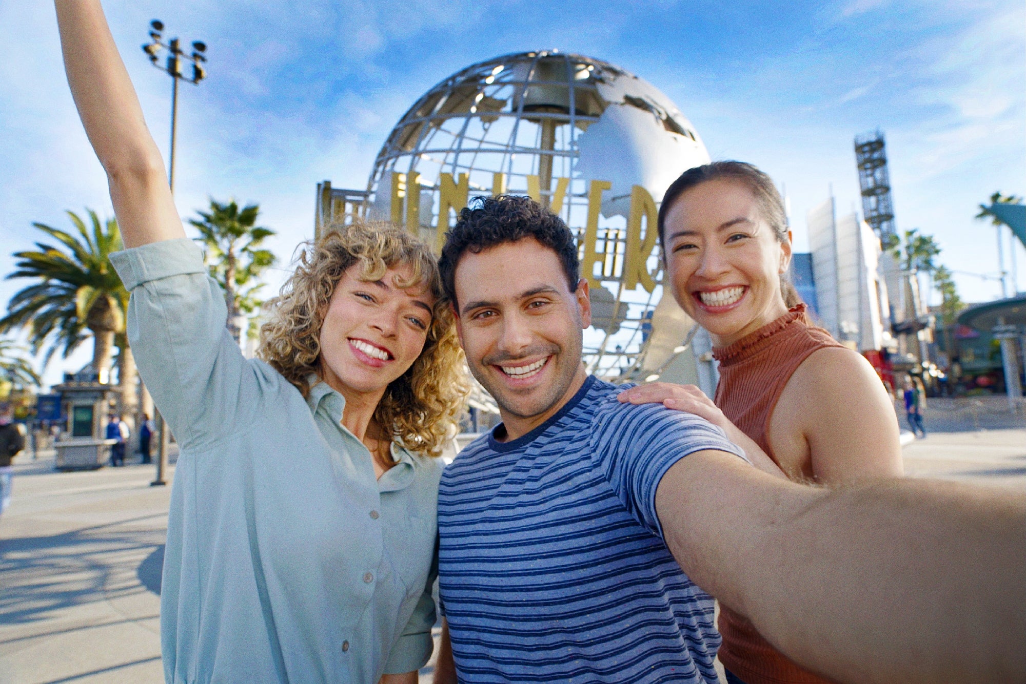 Three people smiling and posing for a selfie in front of the Universal Studios globe sculpture on a sunny day.