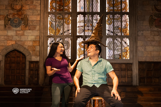 Two people pose in front of large stained glass windows; one wears a pointed hat while sitting on a stool and the other gestures toward them, both smiling.