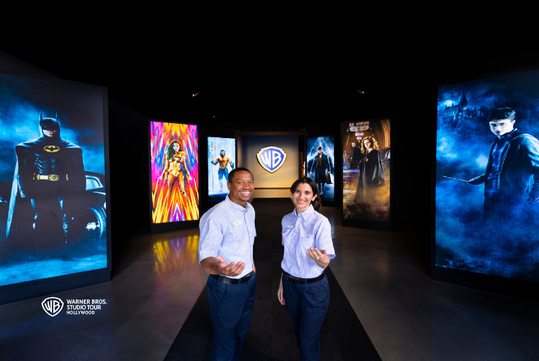 Two Warner Bros. Studio Tour guides stand in front of large illuminated posters of Batman, Wonder Woman, and Harry Potter characters in a dimly lit hallway.