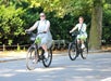 Two people ride bicycles on a sunny day in a park, wearing casual summer clothing and helmets.
