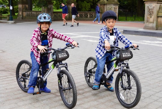Two children wearing helmets and plaid shirts riding bicycles on a paved path in a park.