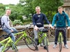 Three young men stand with green bicycles in a park setting.