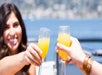 Two people clinking glasses of orange beverage outdoors with water and a blurred cityscape in the background.