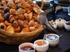 A large basket of assorted pastries and muffins sits on a table next to bowls of various jams and metal tongs.