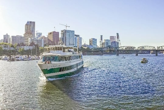 A white and green passenger boat travels on a river with a city skyline and a bridge in the background under a clear sky.