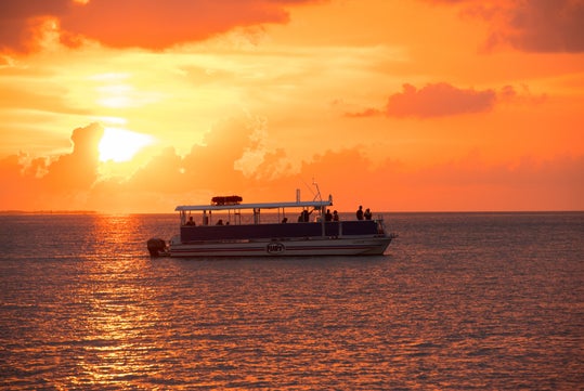 A boat with several people onboard floats on calm water at sunset, with orange and yellow hues filling the sky and reflecting on the sea.