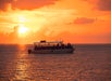 A boat with several people onboard floats on calm water at sunset, with orange and yellow hues filling the sky and reflecting on the sea.