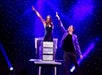 A magician performs an illusion on stage with a woman in a box, both raising one arm, under purple stage lights and a starry backdrop.