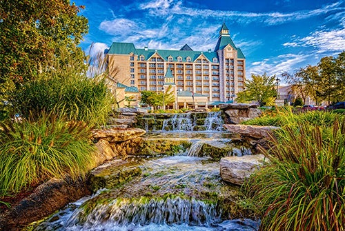Luxurious hotel with green rooftops in the background, a cascading waterfall and lush greenery in the foreground.
