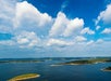 A scenic view of a large lake under a blue sky with scattered white clouds. Green and brown islands are visible in the water, surrounded by distant shoreline.