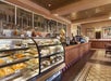 A bakery with glass display cases filled with pastries and cakes. The interior features tiled floors, ceiling details, and a menu board. Two framed posters are on the wall.