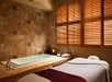 A spa room with a massage table, a lit candle, and a towel beside a tiled bathtub. Wooden shutters cover the window.