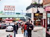 People walk along a street near Pike Place Market in Seattle, with the market’s red sign visible in the background. Some pedestrians appear to be talking and taking photos.