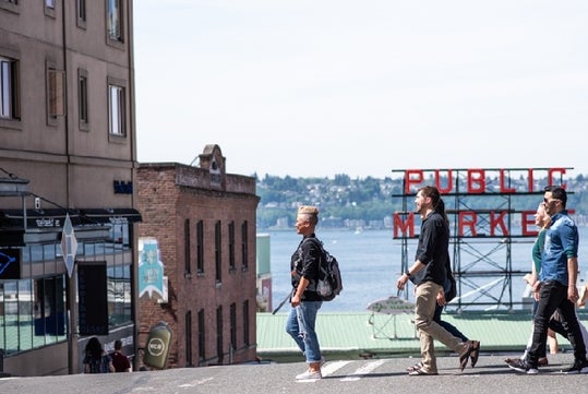 Four people walk across a street with the Public Market sign and waterfront visible in the background on a sunny day.