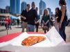 A piece of cooked salmon sits on white paper atop a red table outdoors, with a blurred group of people and city buildings in the background.
