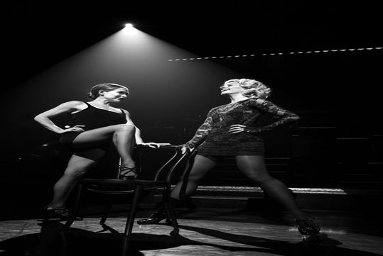 Two performers in elegant outfits strike dynamic poses on stage, with dramatic lighting highlighting their expressions.