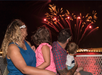 A family of four sits on a bench at night, watching a fireworks display in the sky.
