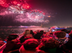 People on a boat watch a fireworks display over the water at night, with bright red and white bursts lighting up the sky.