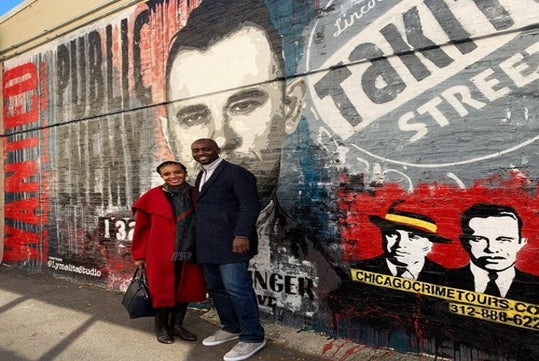 Two people stand smiling in front of a colorful mural depicting historical crime figures and advertising a Chicago crime tour. The mural features faces and text.