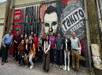 A group of people stands in front of a large mural depicting a bearded man's face with text in the background.
