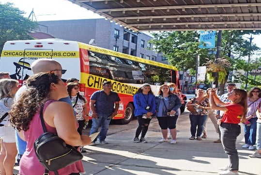 A tour guide speaks to a group of people standing near a red Chicago Crime Tours bus parked on a city street.